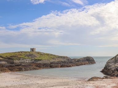 A sandy beach with rocky cliffs and a small stone building on a grassy hill near the sea at Plas Coch Rhoscolyn in Rhoscolyn