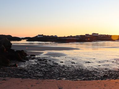A coastal scene at sunset with houses on the shore and rocky beach in the foreground at Plas Coch Rhoscolyn