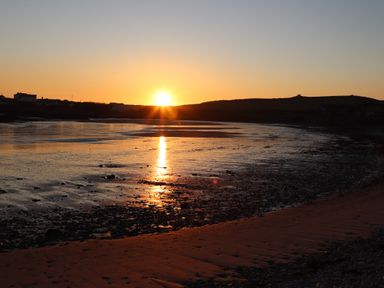 A sunset over a beach with water reflecting the sun and houses on the horizon at Plas Coch Rhoscolyn in Rhoscolyn
