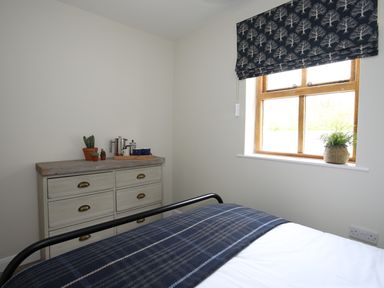 A bedroom with a bed covered in a blue plaid blanket a cream dresser with plants and coffee items and a window with leafy patterned blind at Plas Coch Rhoscolyn in Rhoscolyn