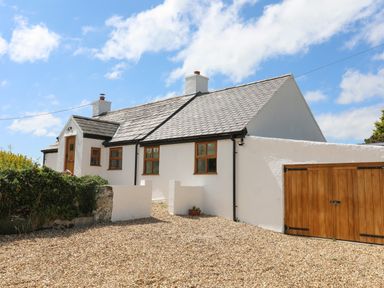 A white cottage with a gray tiled roof and wooden doors beside a gravel driveway at Plas Coch Rhoscolyn in Rhoscolyn