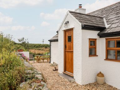 The exterior of a white cottage with a wooden door and windows surrounded by gravel garden with plants and outdoor seating at Plas Coch Rhoscolyn