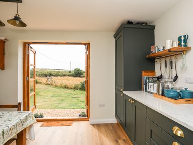 A kitchen with green cabinets and wooden table open door showing a garden at Plas Coch Rhoscolyn