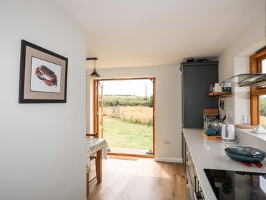 A kitchen with a countertop, cabinets, a table with a cloth, and an open door leading to a grassy outdoor area at Plas Coch Rhoscolyn in Rhoscolyn