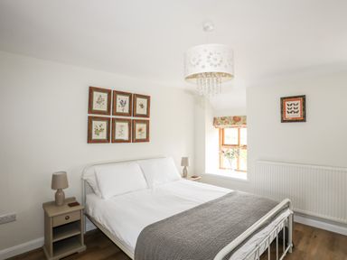 A bedroom with a white metal bed frame bedside tables lamps framed botanical prints a window with floral blinds and a radiator at Plas Coch Rhoscolyn in Rhoscolyn