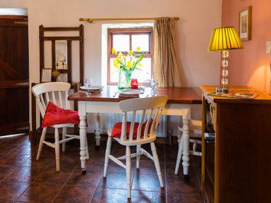 A dining area with a wooden table set with plates glasses and a vase of flowers white chairs with red cushions and a wooden cabinet with a lamp at The Stable in Fethard-On-Sea County Wexford