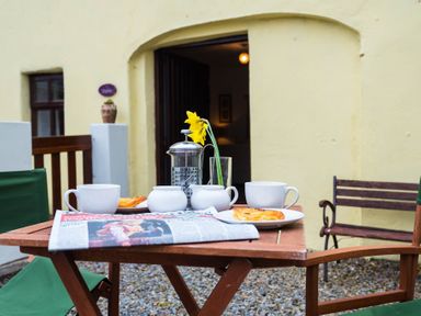 An outdoor wooden table with cups plates pastries a newspaper a French press and a yellow flower in a glass at The Stable in Fethard-On-Sea County Wexford