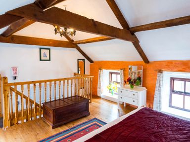 An attic bedroom with wooden beams a wooden chest a white dresser with mirror two windows and a bed with red bedding at The Stable in Fethard-On-Sea County Wexford
