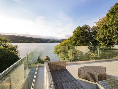 An outdoor patio with wicker furniture and glass railing overlooking a lake and trees at Kessock in Bowness-On-Windermere