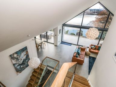 A modern living room with brown leather sofas, turquoise cushions, large windows overlooking water, and a wooden staircase at Kessock in Bowness-On-Windermere