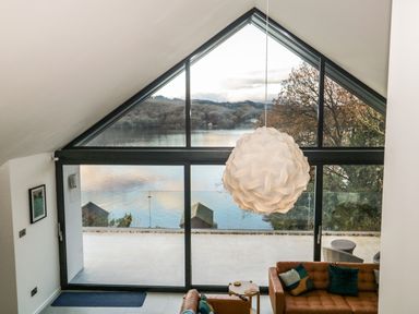 A living room with a large triangular window overlooking a lake with hills and trees at Kessock in Bowness-On-Windermere