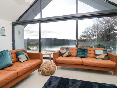 A living room with brown leather sofas and decorative pillows by large glass windows overlooking a lake at Kessock in Bowness-On-Windermere