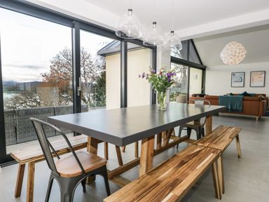 A dining area with a table, benches, metal chairs, and a vase with flowers next to large glass windows at Kessock in Bowness-On-Windermere