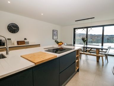 A kitchen with an island countertop a sink and a stove a dining table with benches and chairs near large windows overlooking a lake at Kessock in Bowness-On-Windermere