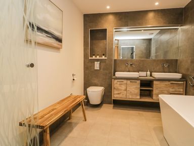 A bathroom with a wooden bench, double sinks on a wooden vanity, a wall-mounted toilet, and a bathtub at Kessock in Bowness-On-Windermere