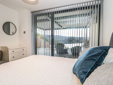 A bedroom with bed pillows a dresser and glass sliding doors with vertical blinds opening to a patio with chairs at Kessock in Bowness-On-Windermere