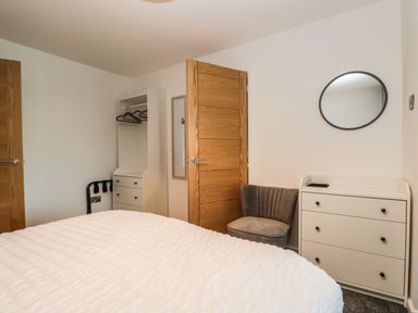A bedroom with a bed covered by a white textured duvet a wooden door a white dresser with three drawers and a round mirror above it a gray upholstered chair and a white open wardrobe with hangers at Kessock in Bowness-On-Windermere