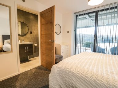 A bedroom with a bed a chest of drawers an open door showing a bathroom and large windows with vertical blinds at Kessock in Bowness-On-Windermere