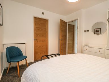 A bedroom with a white bedspread wooden doors a teal chair and a white dresser with a round mirror at Kessock in Bowness-On-Windermere