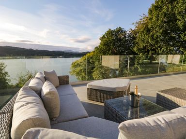 A balcony with a wicker sofa set glass table with a champagne bottle and glass overlooking a lake and trees at Kessock in Bowness-On-Windermere