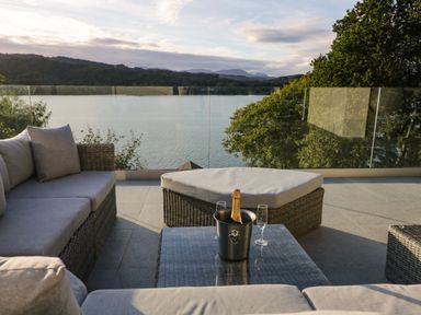 An outdoor seating area with wicker furniture cushions a glass table with a champagne bucket and glasses overlooking a lake and trees at Kessock in Bowness-On-Windermere