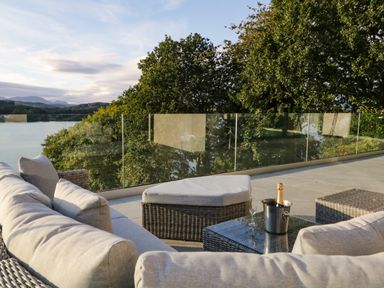 An outdoor seating area with a cushioned sofa, ottoman, glass table with a bottle in an ice bucket and glass on a terrace with glass railing overlooking trees and water at Kessock in Bowness-On-Windermere