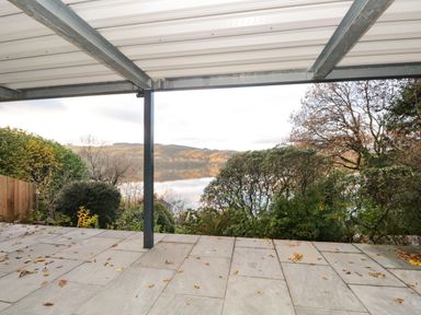 A covered patio with stone tiles surrounded by bushes and trees overlooking a lake and hills in the background at Kessock in Bowness-On-Windermere