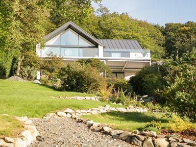 A modern house with large triangular windows surrounded by trees and shrubs with a stone pathway leading to it at Kessock in Bowness-On-Windermere