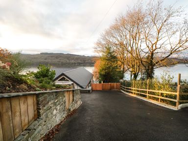 A paved driveway with a stone and wood retaining wall leading to a house near a body of water with trees in the background at Kessock in Bowness-On-Windermere