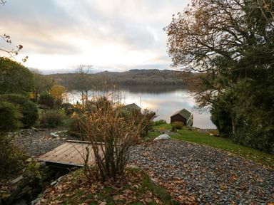 A lakeside garden with a small wooden bridge shrubs and boathouses by the water at Kessock in Bowness-On-Windermere