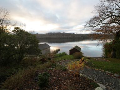 A garden with trees shrubs and a pathway leading to two boathouses by a lake at Kessock in Bowness-On-Windermere