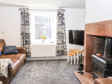 A living room with a brown leather sofa a window with patterned curtains a fireplace with a wood stove a flat screen tv and nesting tables at Sparrow Cottage in Keswick