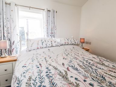 A bedroom with a bed with floral bedding and pillows two bedside tables with lamps and a window with floral curtains at Sparrow Cottage in Keswick