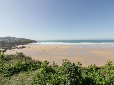 A sandy beach with waves and green shrubs in the foreground at Fistral Apres Surf in Newquay