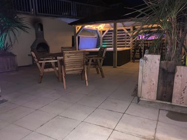 An outdoor patio with a wooden table and chairs a chimney and a covered hot tub with string lights at Fistral Apres Surf in Newquay