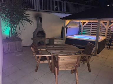 An outdoor patio with a round wooden table and four chairs a hot tub with a wooden pergola a built in barbecue grill and a potted plant at Fistral Apres Surf in Newquay