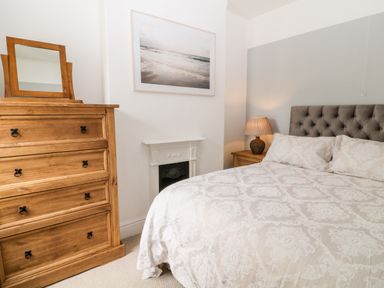 A bedroom with a bed with patterned bedding a wooden chest of drawers with a mirror a bedside table with a lamp and a framed photo above a fireplace at Daisy Cottage in Bideford
