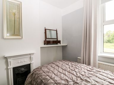 A bedroom corner with a bed covered in a textured duvet a small decorative fireplace a wall shelf with a mirror a window with curtains and a framed floral artwork at Daisy Cottage in Bideford