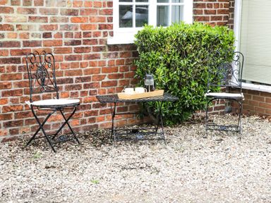 A small outdoor seating area with two metal chairs and a metal table with a tray holding a French press and cups by a brick wall and window at Chestnut Cottage in Filey