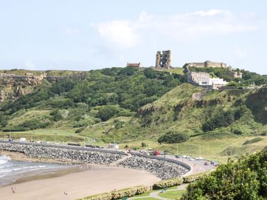 A coastal road with cars and a beach below green hills with castle ruins at Chestnut Cottage in Filey