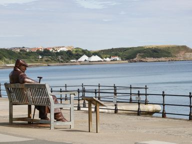 A seaside promenade with a metal sculpture of a seated person on a bench overlooking the water