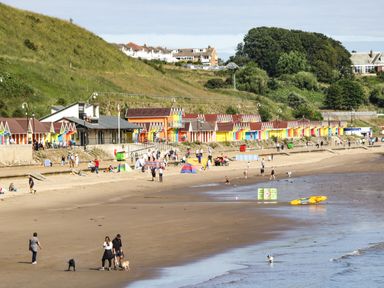 A beach with people walking dogs near colorful beach huts and grassy hills at Chestnut Cottage in Filey