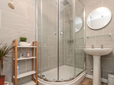 A bathroom with a curved glass shower enclosure a white pedestal sink a round mirror and a small wooden shelf with toiletries at Chestnut Cottage in Filey