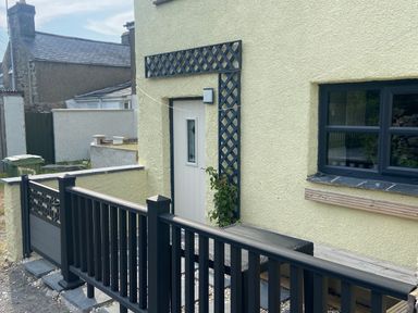 The exterior of a house with a light-colored door, a black fence, a window, and a climbing plant at Weaver's Cottage in Tremadog