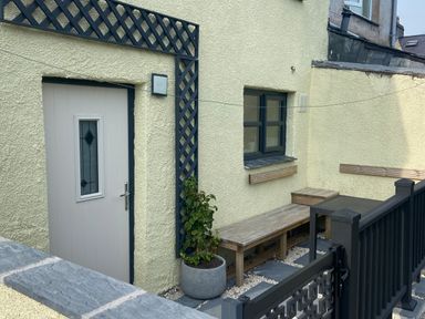 An outdoor seating area with a wooden door a small window benches a potted plant and a black fence at Weavers Cottage in Tremadog