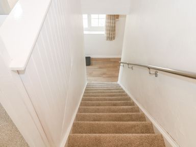 A carpeted staircase with white walls and a metal handrail leading to a wood floor hallway with a window at Rose Cottage in Marldon
