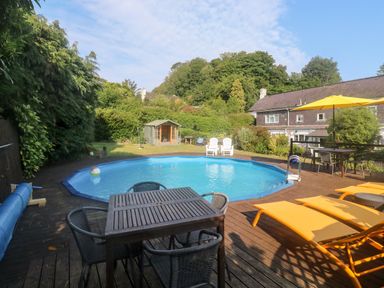 An outdoor area with a swimming pool surrounded by wooden decking tables chairs sun loungers and a yellow umbrella at Rose Cottage in Marldon
