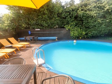 An outdoor pool with blue water surrounded by wooden deck chairs tables and green trees at Rose Cottage in Marldon