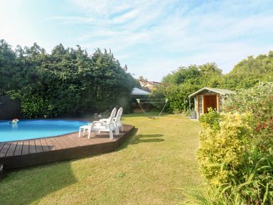 A backyard with a small round pool and two white chairs on a wooden deck a swing set on grass and a wooden garden shed surrounded by bushes