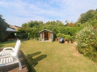 A garden with a small shed a badminton net white plastic chairs and plants at Rose Cottage in Marldon